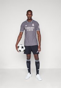 Maglia da calcio grigia con texture trapuntata e dettagli bianchi, pantaloni corti neri, calzettoni neri con strisce, che tiene un pallone da calcio bianco e nero.