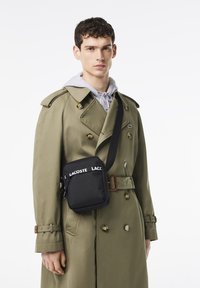 Borsa a tracolla nera in tessuto con cinturino con la scritta "LACOSTE", indossata da un modello con un hoodie grigio chiaro e un trench beige.