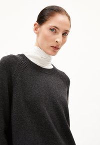 Femme avec les cheveux gominés, portant un pull gris foncé par-dessus un col roulé blanc, regardant directement l'objectif contre un fond blanc.
