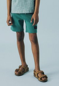 Tealfärgade shorts med sidofickor och svart etikett, kombinerade med bruna sandaler med remmar som har en rosett och en kilklack.