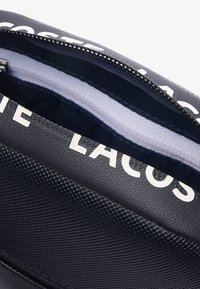 Interior de bolsa negra texturizada con un zipper blanco, con un forro azul claro. El exterior muestra letras blancas prominentes.