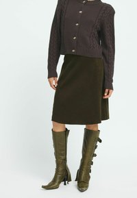 Brauner, kabelgemusterter, verkürzter Pullover, olivgrüner Cordrock im A-Linien-Schnitt und knielange olivgrüne Lederstiefel mit Schnallenakzenten.