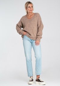 Kobieta stoi z ręką w kieszeni, ubrana w oversize'owy brązowy sweter, jasnoniebieskie dżinsy, trampki w panterkę i złote kolczyki w kształcie kół.