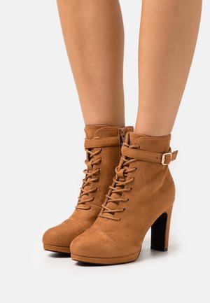 Bottines en suède beige avec un talon haut, un devant lacé, une fermeture éclair sur le côté et une sangle à boucle autour de la cheville. Bout rond.