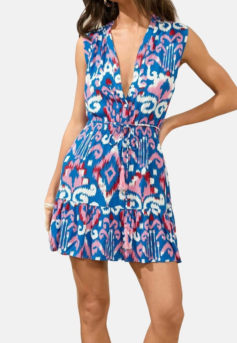 Femme portant une robe bleue sans manches avec des motifs abstraits roses, blancs et rouges, un décolleté en V profond et une ceinture nouée à la taille.