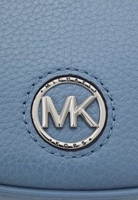 Sac en cuir bleu clair avec une surface texturée. Présente un logo circulaire argenté avec les initiales "MK" et des coutures contrastantes.