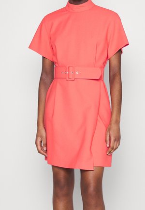 Woman wearing short-sleeve coral dress with high neck and matching wide belt, standing against plain light background.