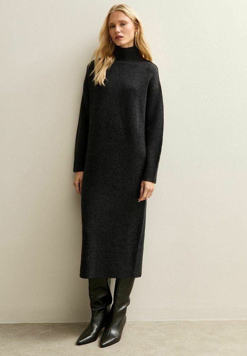 Abito nero in maglia con collo alto, maniche lunghe e orlo a coste. Abbinato a stivali neri a punta alti fino al ginocchio. Design minimalista.