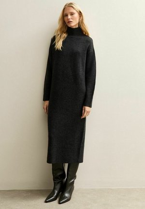 Vestido negro de punto con cuello alto, mangas largas y dobladillo acanalado. Combinado con botas de tacón alto negras con punta. Diseño minimalista.