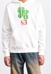 Person klädd i en vit hoodie med gröna monsterablad och röd Nike-logotyp, tillsammans med blå gradiente jeans, som står framför en enfärgad bakgrund.