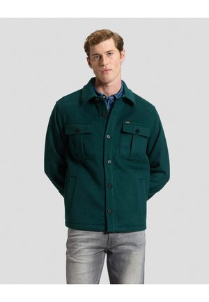 Junger Mann mit kurzen lockigen Haaren trägt eine dunkelgrüne, geknöpfte Jacke über einem blauen Hemd und hellgrauen Jeans, steht vor einem schlichten Hintergrund.