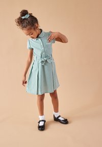 Groen-witte geruite jurk met korte mouwen, knopen aan de voorkant, tailleband en geplooide rok. Draagt zwarte schoenen en witte enkelsokken.