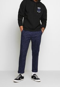 Černá mikina s kapucí a přední kapsou, s logem, doplněná o kalhoty s námořnickým leopardím potiskem a černé tenisky s bílou podrážkou a kotníkovými ponožkami.