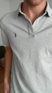 Pruhované polo tričko sivé a biele vyrobené z bavlny, so stojatým golierom a malým logom na ľavej strane. Viditeľný dvojitý gombíkový zapínanie.