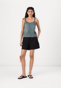 Denim top bez rukava s podesivim vezicama, u izblijedjeloj plavoj boji, upareno s crnim šortsevima. Model drži malu bež torbicu i nosi crne japanke.