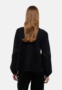 Blouse noire avec des manches bouffantes, des accents en dentelle transparente et une coupe décontractée. Texture lisse avec un léger brillant.