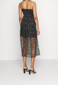 Robe noire avec un superposé transparent à imprimé léopard et des bretelles spaghetti, dotée d'un corsage ajusté et d'une jupe fluide de longueur mi-mollet. Sandales noires à talons blocs.