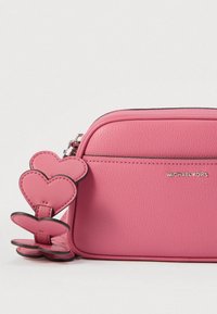 Ružičasta kožna torba glatke teksture, s patentnim zatvaračem i ukrasnim srcastim privjescima pričvršćenima za zatvarač. Vidljiv naziv marke.