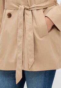 Beige beltejakke med knappelukking, glatt tekstur, sidelommer og skreddersydd passform, kombinert med blå jeans.