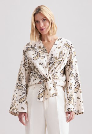 Blouse crème à motif cachemire avec un design croisé, manches longues et un nœud à l'avant. Présente des accents noirs et dorés ainsi qu'une texture lisse.