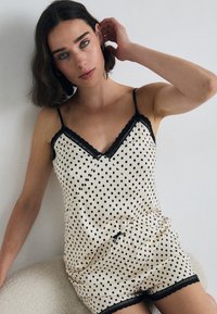 Svart polkadott satin søvnsett med blonderdetaljer, som inneholder en tettsittende tank top og shorts. Fargen er kremhvit med svarte detaljer.