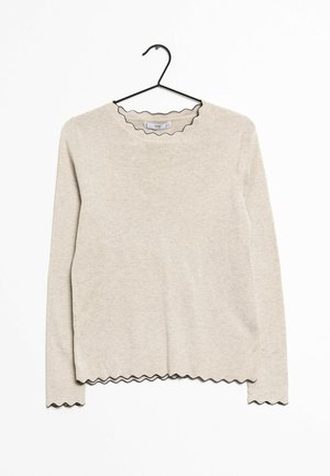 Beige langwerpige trui met lange mouwen en een geribbelde textuur, voorzien van een geschulpte zwarte afwerking rond de kraag en de zoom. Hangt aan een zwarte haak.