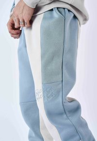 Pantalones de chándal azul claro y blancos con diseño en bloques de color, textura suave y detalle bordado en la pierna. Incluyen cinturilla elástica.