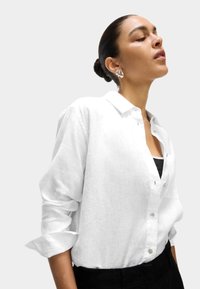 Camisa de lino blanca con mangas largas y diseño abotonado. Presenta un cuello clásico y una textura sutil. Se lleva con una camiseta negra debajo.