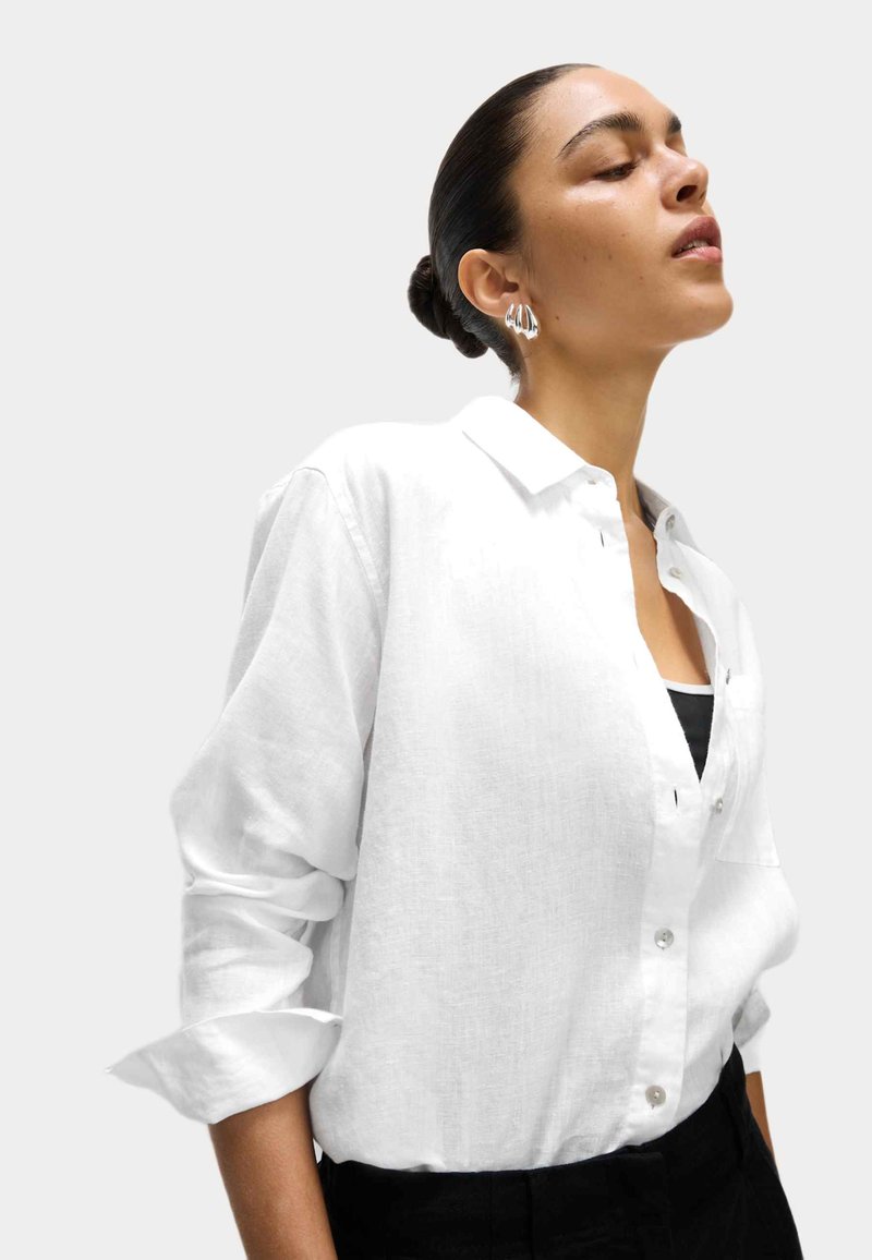 Camisa de lino blanca con mangas largas y diseño abotonado. Presenta un cuello clásico y una textura sutil. Se lleva con una camiseta negra debajo.