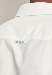 Biała koszula z kołnierzykiem, z niebieską metką "JOULES" wszytą z tyłu. Gładka faktura, regularny krój.