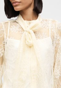 Μπλούζα από δαντέλα σε κρεμ χρώμα με φιόγκο στη λαιμόκοψ η. Ανθικά μοτίβα στις διαφανείς μανίκες, με τετράγωνη λαιμόκοψη και διακοσμητικό κουμπί.