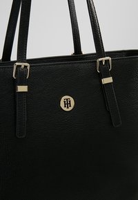 Sac fourre-tout en cuir noir avec une finition texturée, des garnitures dorées et un logo circulaire. Dispose de deux poignées et d'un design structuré.