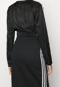 Person klädd i en svart långärmad tröja och svart kjol med elastisk midja och vita vertikala ränder på sidorna, visad bakifrån.