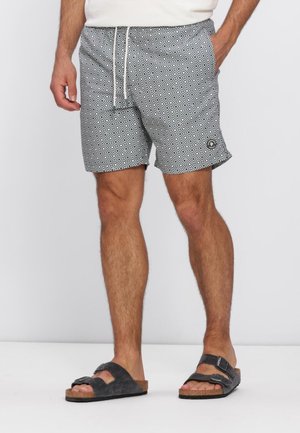 Man draagt zwart-wit geruit korte broek met trekkoord en zwarte sandalen met dubbele band, staand op een witte vloer.