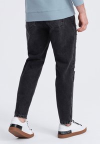 Jeans de mezclilla negra con un corte relajado, que presentan un detalle desgastado en la pierna, combinados con zapatillas blancas y un sudadera azul.