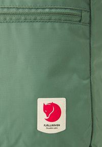 Borsa in tessuto verde con zip, caratterizzata da una patch bianca con il logo di una volpe rossa e il testo "FJÄLLRÄVEN Sweden 1960." Materiale testurizzato e resistente.