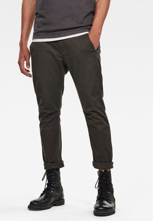 Mężczyzna w ciemnooliwkowych spodniach podwiniętych do góry i czarnych skórzanych butach sznurowanych, stojący z jedną ręką w kieszeni na białym tle.