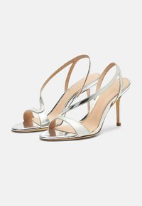 Srebrne metalne sandale s visokim potpeticama, otvorenih prstiju, tanke trake za strap-on i stiletto potpetice na bijeloj pozadini.