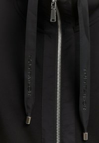 Zwarte jas met ritssluiting en zilverkleurige ritssluiting, voorzien van zwarte trekkoorden met merkopdruk en een gladde stoftextuur.