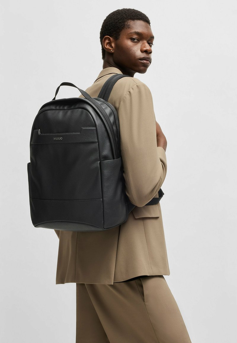 Sac à dos en cuir noir avec une finition texturée, une forme contournée, une poche avant et une marque discrète. Bandoulières adjustables et construction robuste.