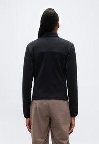 Άτομο με μακριά μαύρα μαλλιά, φορώντας μαύρο σακάκι και μπεζ παντελόνι, στέκεται με την πλάτη προς το μέρος μας, μπροστά σε ένα απλό ανοιχτόχρωμο φόντο.