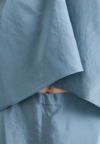 Lys blå stofftop med drapert design, som har en teksturert overflate. En eksponert midje viser et gullsirkelformet smykke.