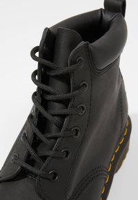 Botín de cuero negro hasta el tobillo con punta redondeada, diseño de cordones, cuello acolchado y costura amarilla a lo largo de la suela.