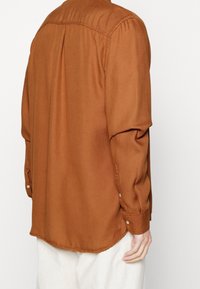 Camicia marrone a maniche lunghe realizzata in un tessuto morbido. Presenta un orlo arrotondato, polsini con bottone e una piega sul retro per un tocco di dettaglio.