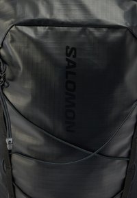 Μαύρο σακίδιο με υφή, με το λογότυπο «SALOMON» να ξεχωρίζει. Διαθέτει κλείσιμο με φερμουάρ και ελαστικά κορδόνια bungee ως διακοσμητικά στοιχεία.