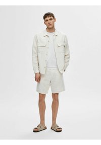 Lichte linnen jas met twee voorzakken, gecombineerd met bijpassende shorts. De outfit wordt compleet gemaakt met beige sandalen. Neutrale kleuren door het geheel.