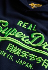 Sudadera marina con bordado verde intenso que dice "REAL SuperDry" y texto en japonés, acentuada por un diseño circular y un tejido texturizado.