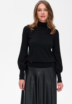 Kobieta z włosami jasno-brązowymi do ramion, nosi czarny sweter z długim rękawem i golfem oraz czarną skórzaną spódnicę, stoi na tle białym.