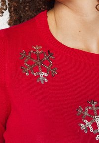 Czerwony sweter z prążkowanym wykończeniem dekoltu i dwoma srebrnymi cekinowymi wzorami płatków śniegu z przodu, prezentujący teksturowany dzianinowy materiał.