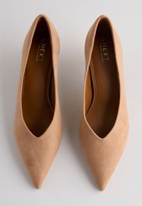 Chaussures à talons hauts en daim couleur nude avec un design à bout pointu. Texture lisse avec une découpe en V profonde, dotées d'une semelle intérieure légèrement rembourrée et d'une quincaillerie minimale.